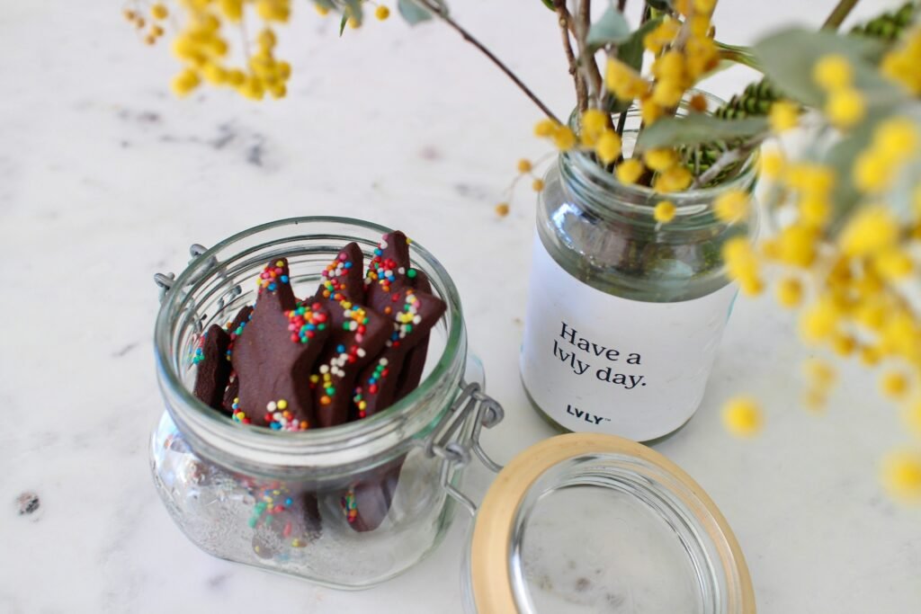 Glass jar filled with star-shaped chocolate biscuits topped with colourful sprinkles, placed beside a floral arrangement in a jar labelled “Have a lvly day” on a white marble surface.